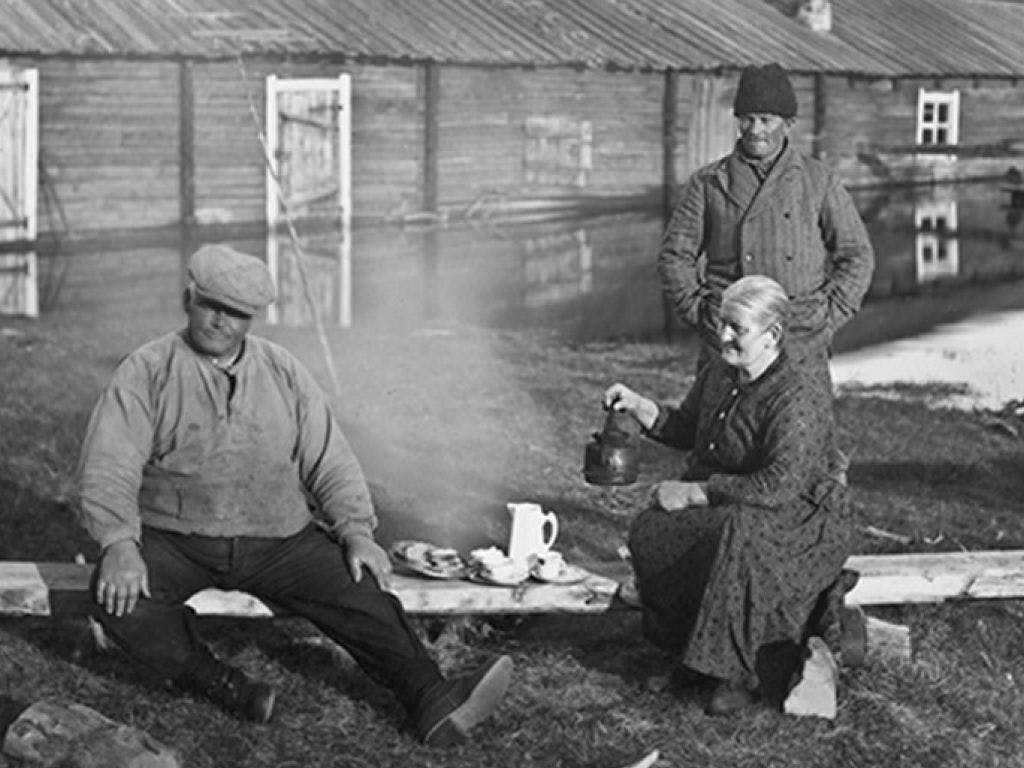 Svart-hvitt bilde av to arbeidere og en gammel kone som skjenker kaffe fra en kaffekjel